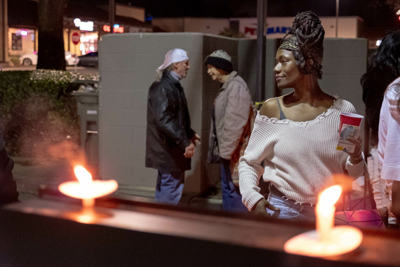 A woman with a headscarf and off-shoulder sweater stands near lit candles at a memorial. In the background, two people converse near a wall.