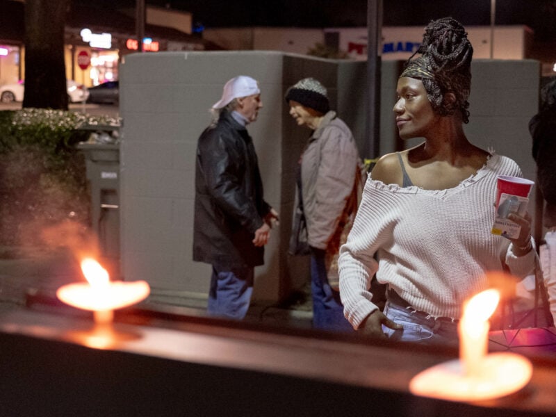 A woman with a headscarf and off-shoulder sweater stands near lit candles at a memorial. In the background, two people converse near a wall.