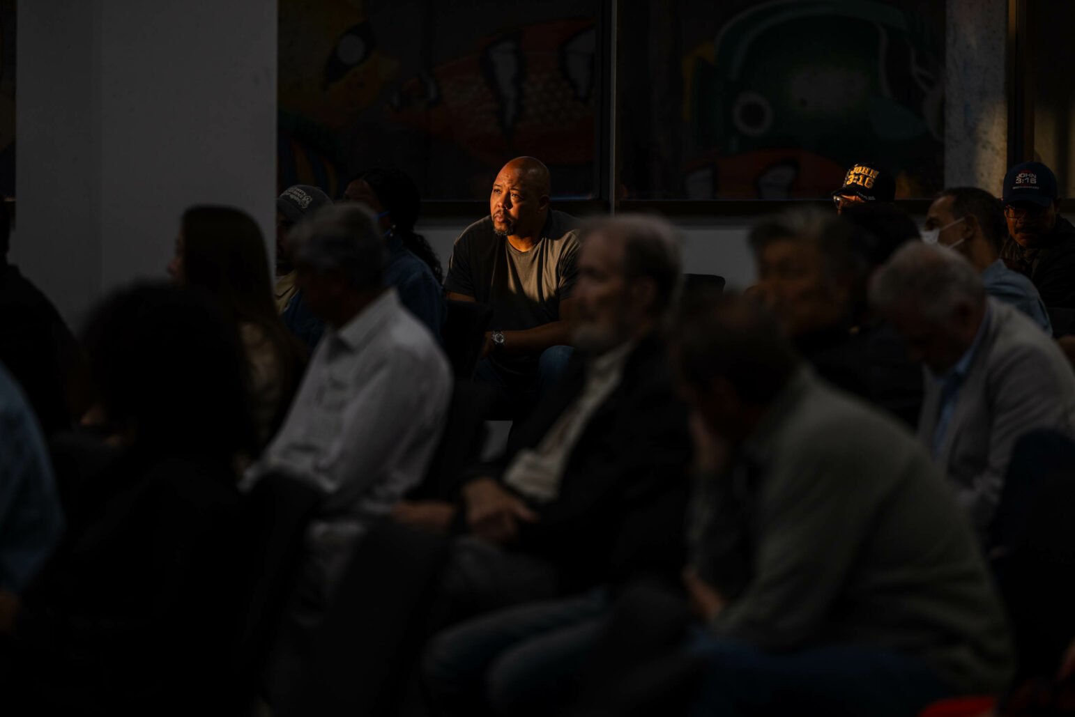 A man bathed in warm sunlight listens to a speaker at a community event while surrounded by a diverse group of community members.