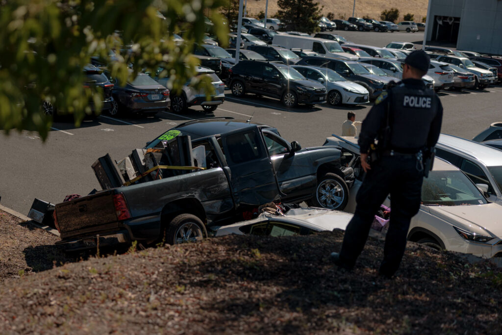 A police officer surveying the crash site from a hillside overlooking the dealership parking lot.
