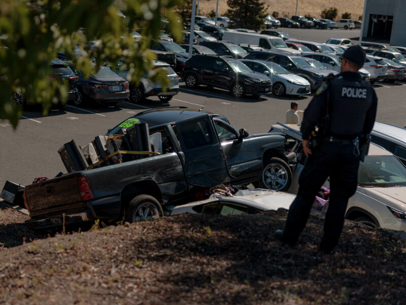 A police officer surveying the crash site from a hillside overlooking the dealership parking lot.