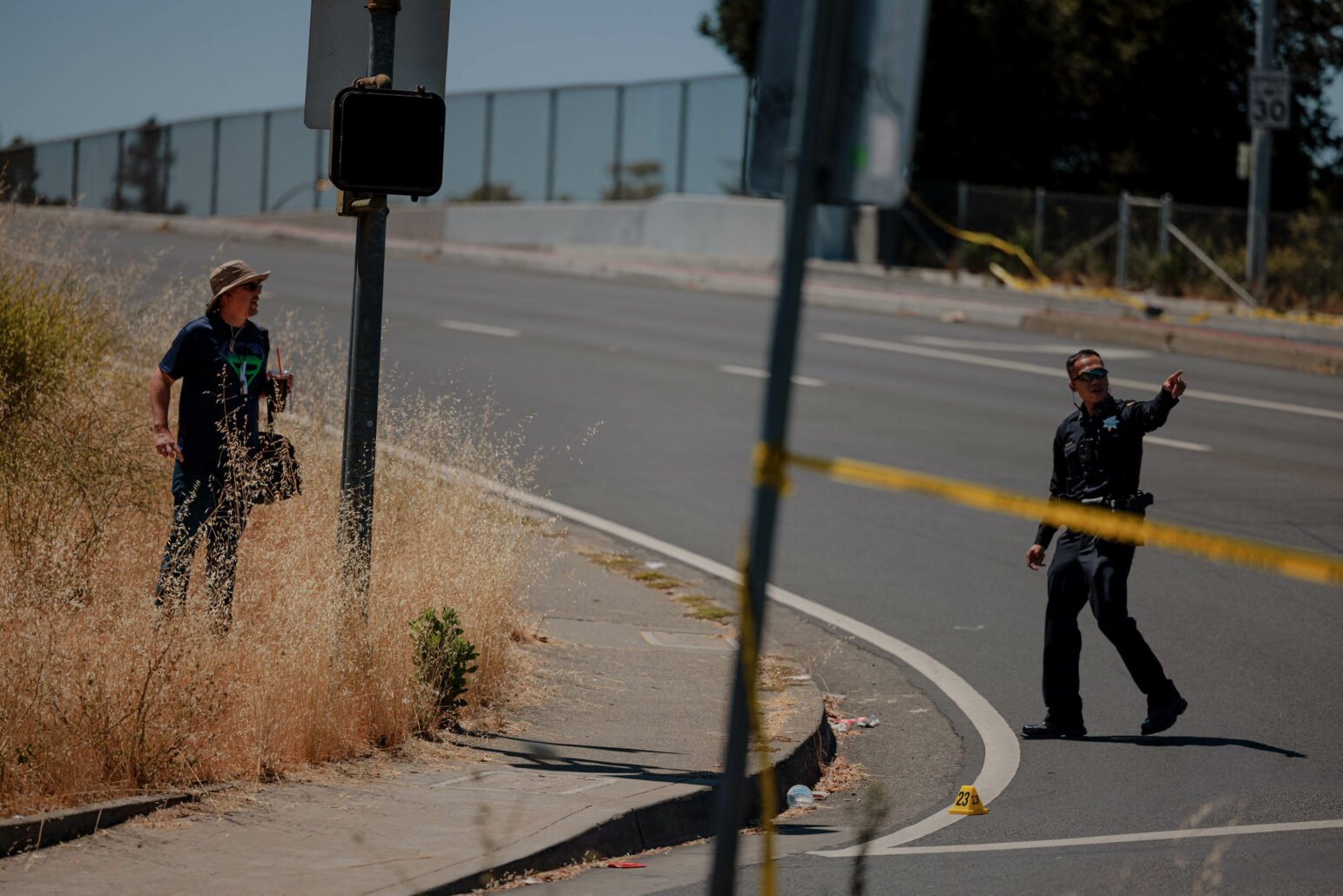 A police officer directs pedestrian traffic at a crime scene. A person with a hat stands nearby on the sidewalk. Yellow police tape and evidence markers are visible, indicating an ongoing investigation.