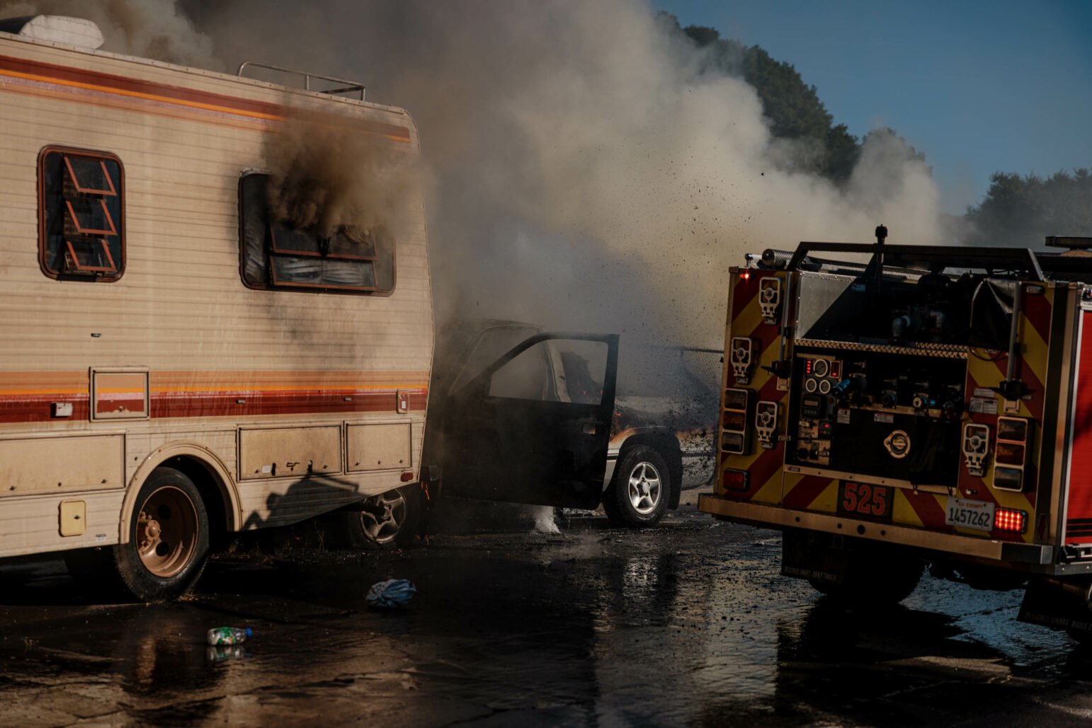 A fire truck and a recreational vehicle (RV) at the scene of a vehicle fire. Smoke billows from the RV and a nearby pickup truck with open doors. Firefighters are on the scene, and water is pooled on the ground.