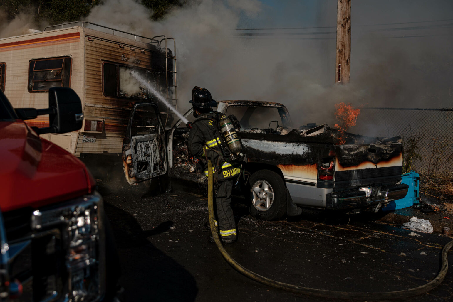 A firefighter in full gear sprays water at a burning vehicle next to an RV. Flames and smoke are visible, and the firefighter works to extinguish the fire. A red fire truck is parked nearby.