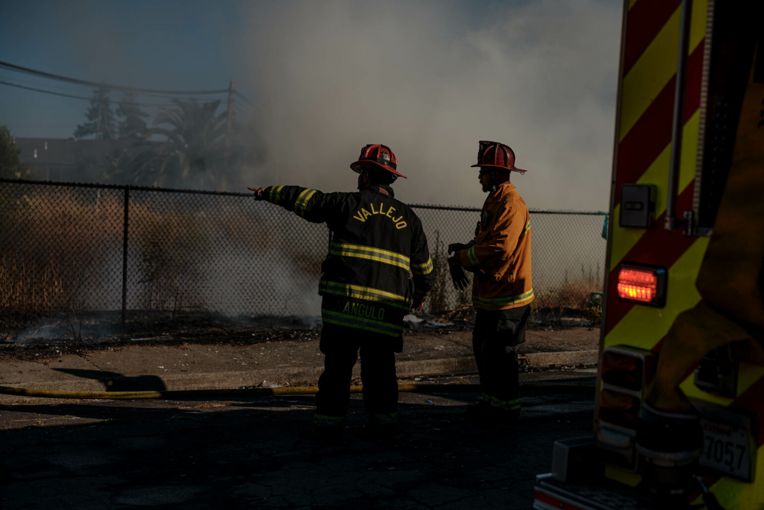 Two firefighters in yellow and black uniforms stand and discuss the situation at the scene of a fire. Smoke rises in the background, and a chain-link fence borders the area.