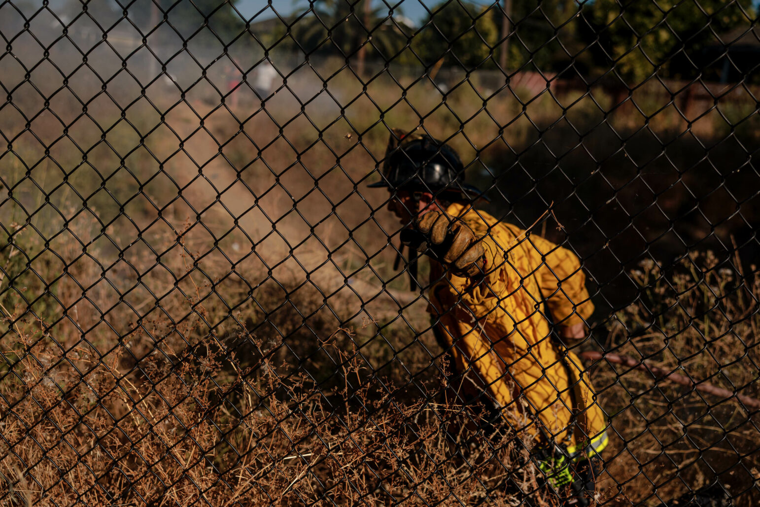 A firefighter in yellow gear pulls a hose along a grassy area behind a chain-link fence. Smoke rises in the background as the firefighter works to control the fire.