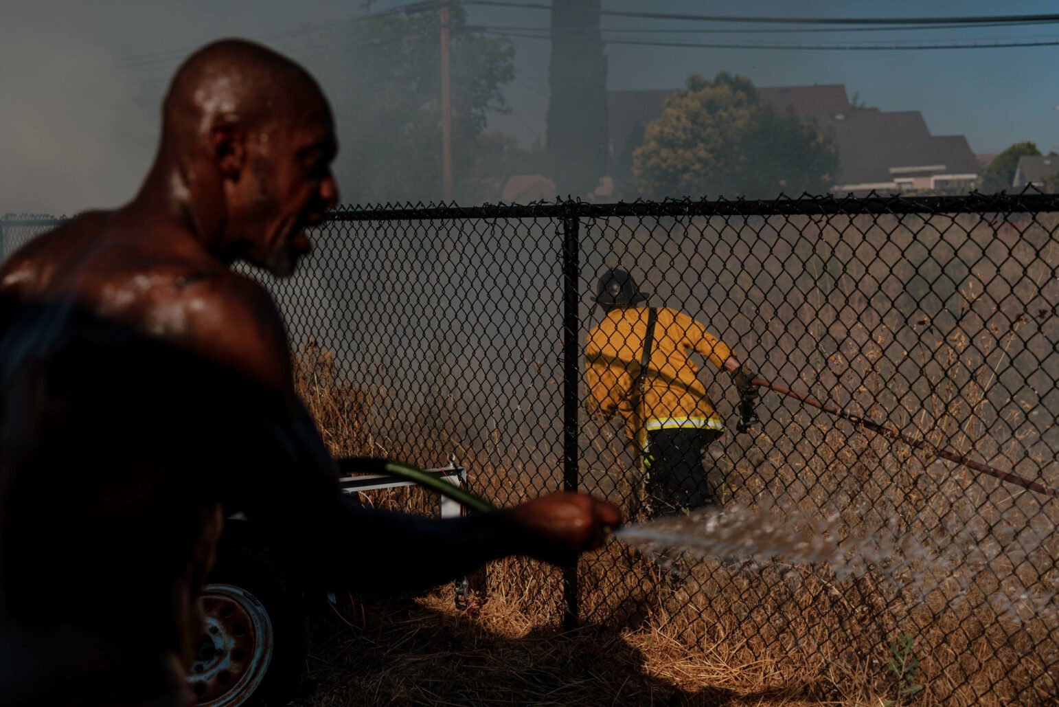 A shirtless man sprays water with a hose while a firefighter in yellow gear works behind a chain-link fence to control a fire in a grassy area. Smoke and flames are visible.