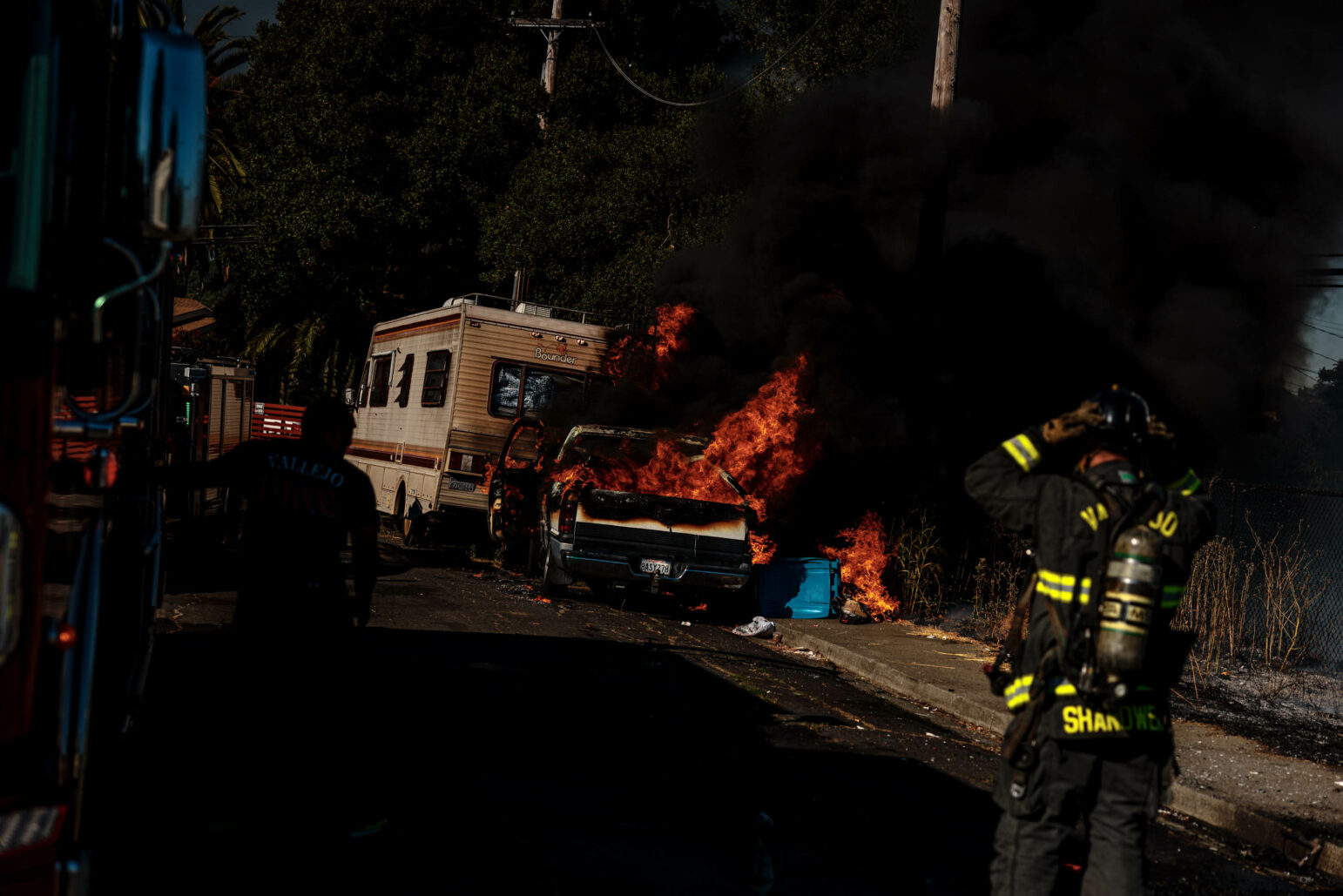 A large vehicle fire with intense flames and black smoke engulfing a car next to an RV. Firefighters in protective gear work to extinguish the blaze, with one firefighter in the foreground adjusting their helmet.