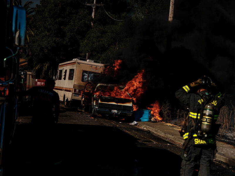 A large vehicle fire with intense flames and black smoke engulfing a car next to an RV. Firefighters in protective gear work to extinguish the blaze, with one firefighter in the foreground adjusting their helmet.