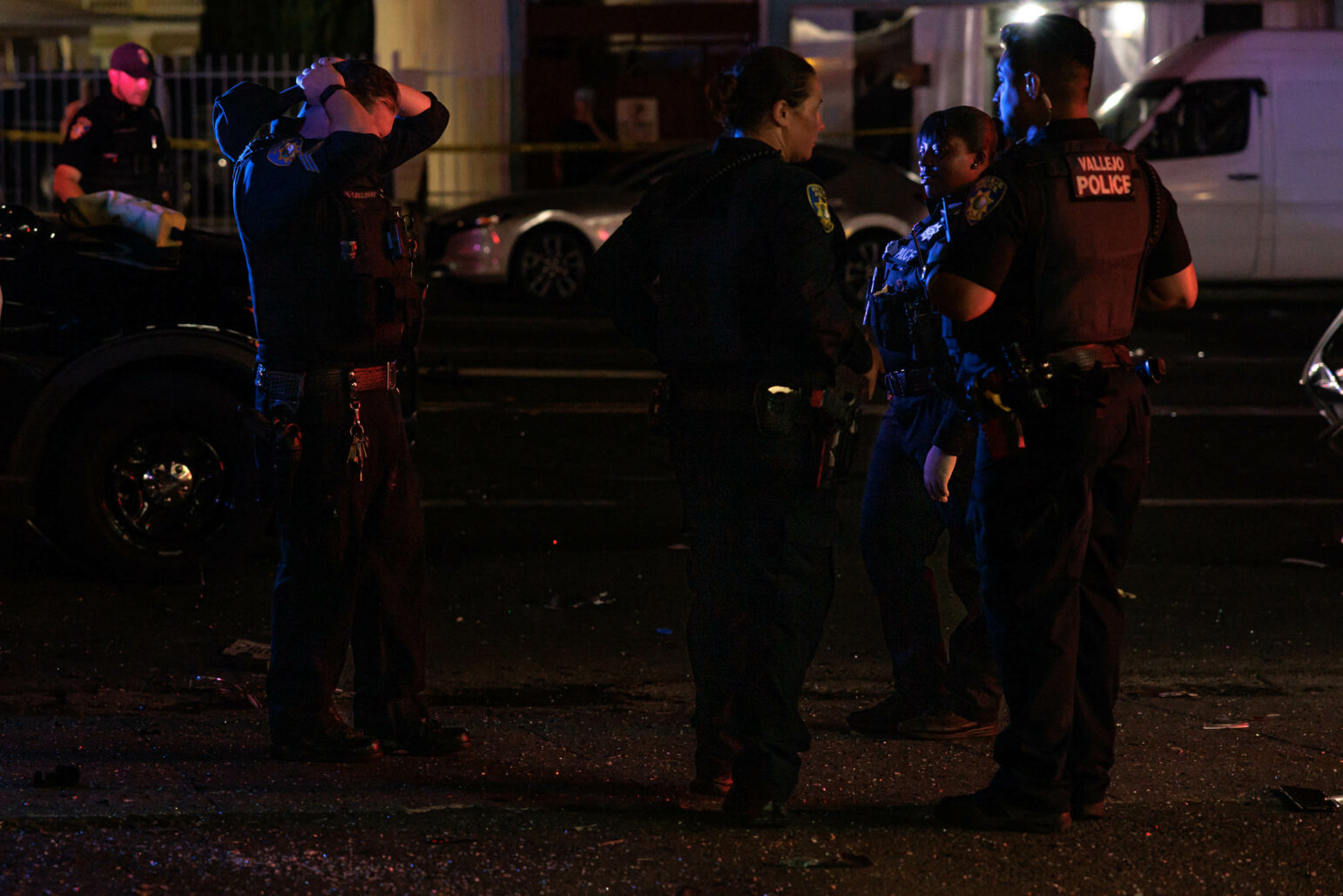 Several police officers gather and converse at the scene of a nighttime collision. One officer is holding their head, while others are engaged in discussion. Debris is scattered on the road, and police tape is visible in the background."