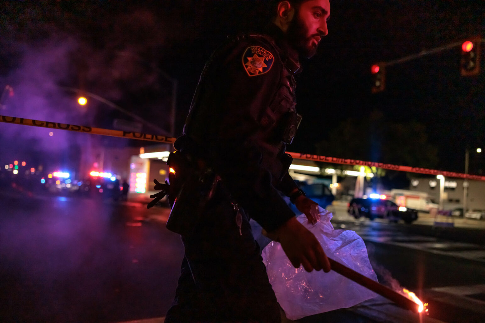 A police officer holding a flare and a plastic bag is seen at the scene of a nighttime collision. Police tape and flashing lights from police cars are visible in the background, with a gas station in the distance.