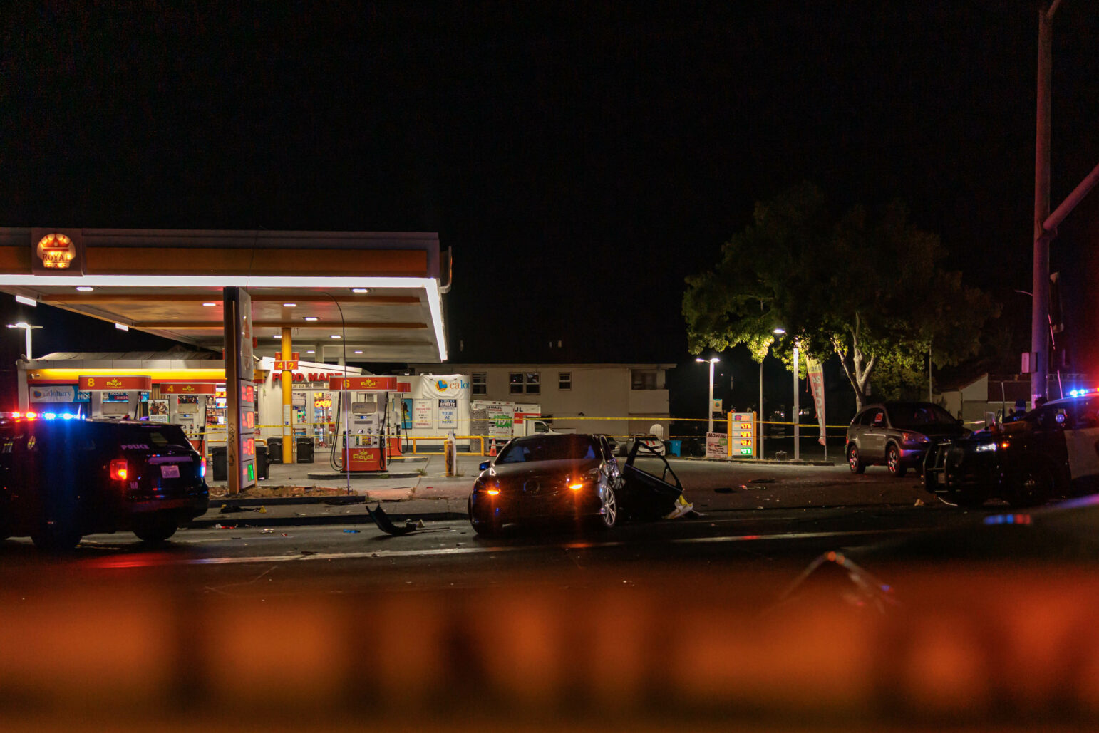 A nighttime scene of a gas station cordoned off by police tape. Multiple vehicles, including police cars with flashing lights, are present at the scene of a collision. Debris is scattered across the road, and the gas station's illuminated signs and fuel pumps are visible in the background.