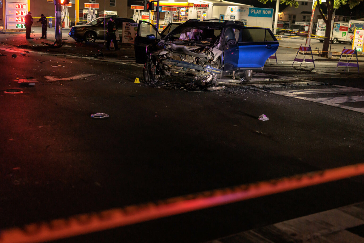 A car with severe front-end damage is seen at night in the middle of an intersection, with debris scattered around. Police officers and emergency responders are at the scene. A gas station is visible in the background with illuminated signs and barricades set up around the area.