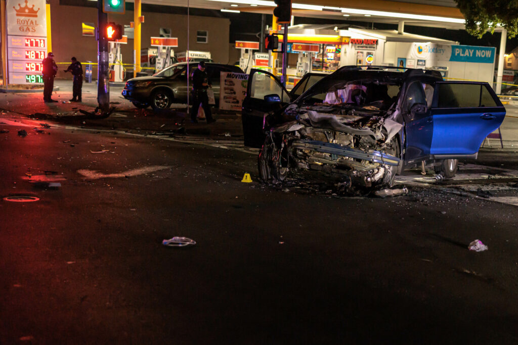 A car with severe front-end damage is seen at night in the middle of an intersection, with debris scattered around. Police officers and emergency responders are at the scene. A gas station is visible in the background with illuminated signs and barricades set up around the area.