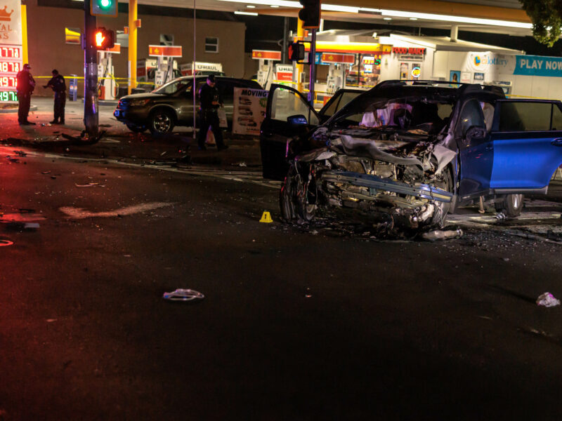 A car with severe front-end damage is seen at night in the middle of an intersection, with debris scattered around. Police officers and emergency responders are at the scene. A gas station is visible in the background with illuminated signs and barricades set up around the area.
