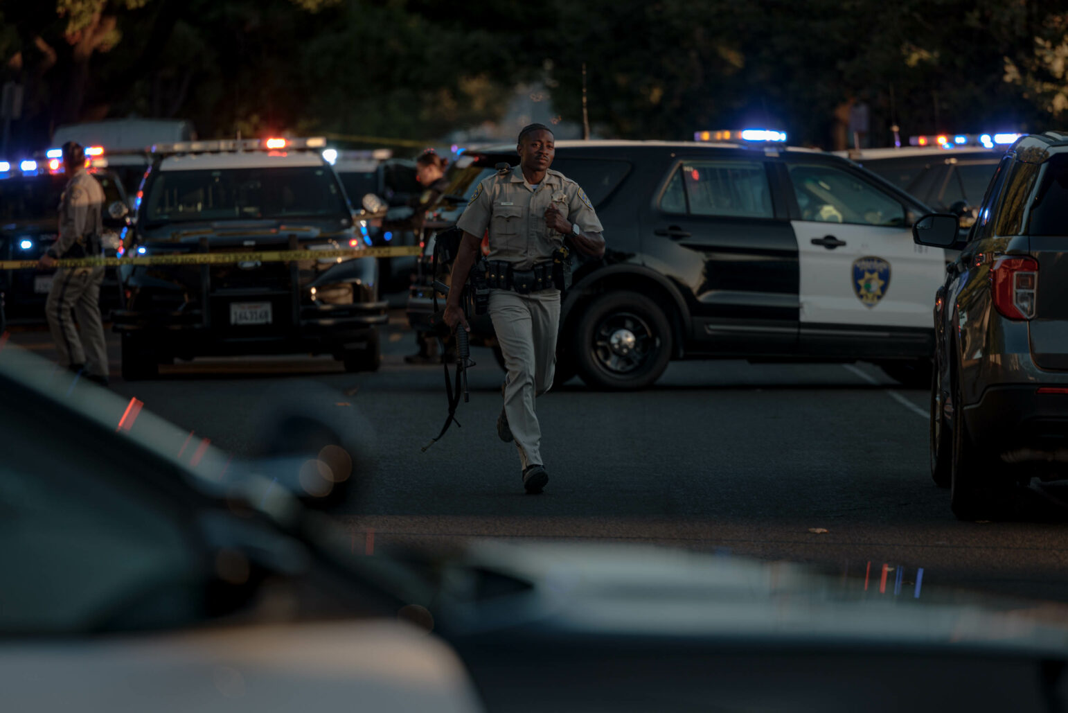 A California Highway Patrol officer holding a rifle runs toward the camera with police vehicles in the background.