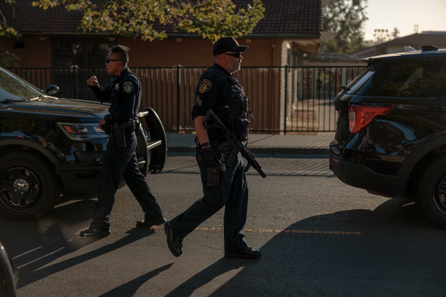 Two police officers in uniform walk beside their patrol cars in the afternoon. One officer is holding a rifle while the other walks with a hand near their mouth. A fence and a building are visible in the background.