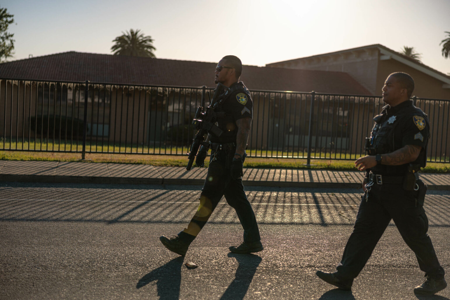 Two police officers in uniform walk along a street in the afternoon. They are carrying rifles, and a fence and a building with a red roof are visible in the background.