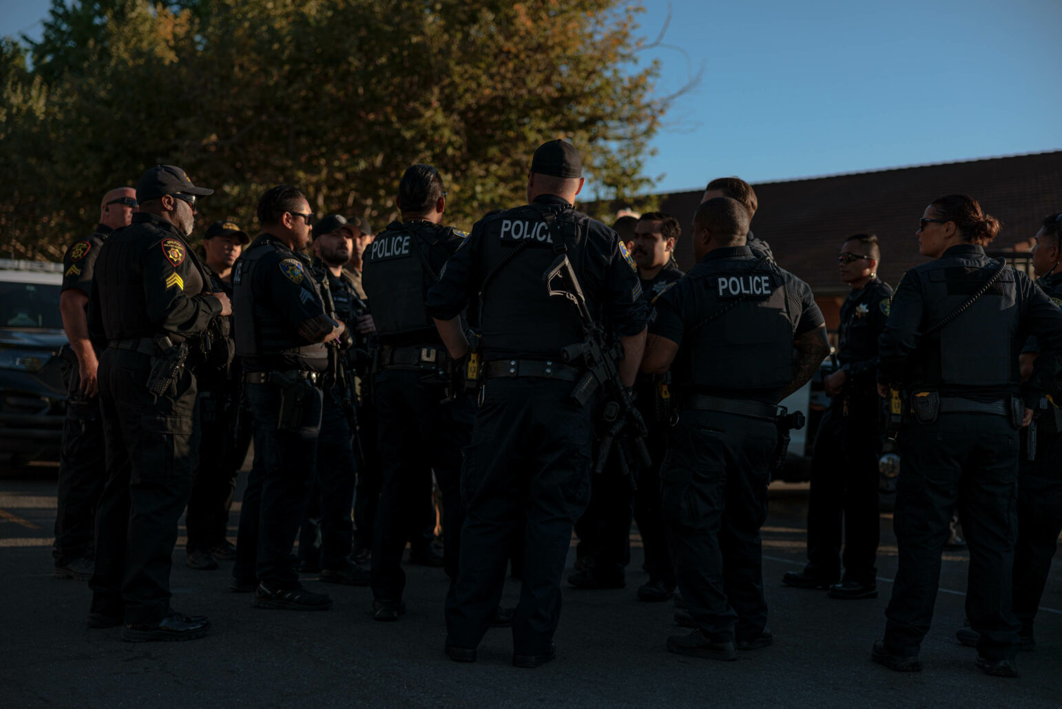 Two police officers in uniform walk along a street in the afternoon. They are carrying rifles, and a fence and a building with a red roof are visible in the background.