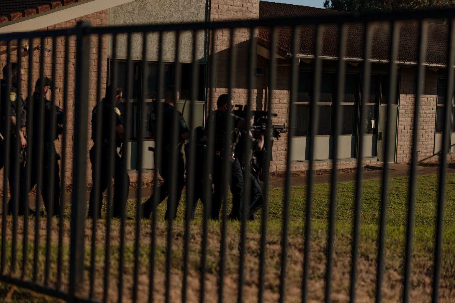 A group of police officers in uniform walk beside a fenced area of a building. They are carrying rifles and other tactical gear.