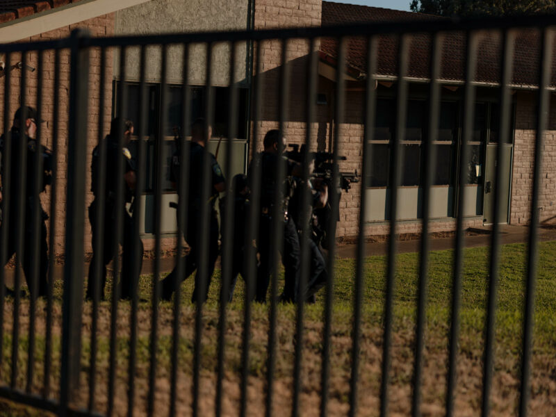 A group of police officers in uniform walk beside a fenced area of a building. They are carrying rifles and other tactical gear.
