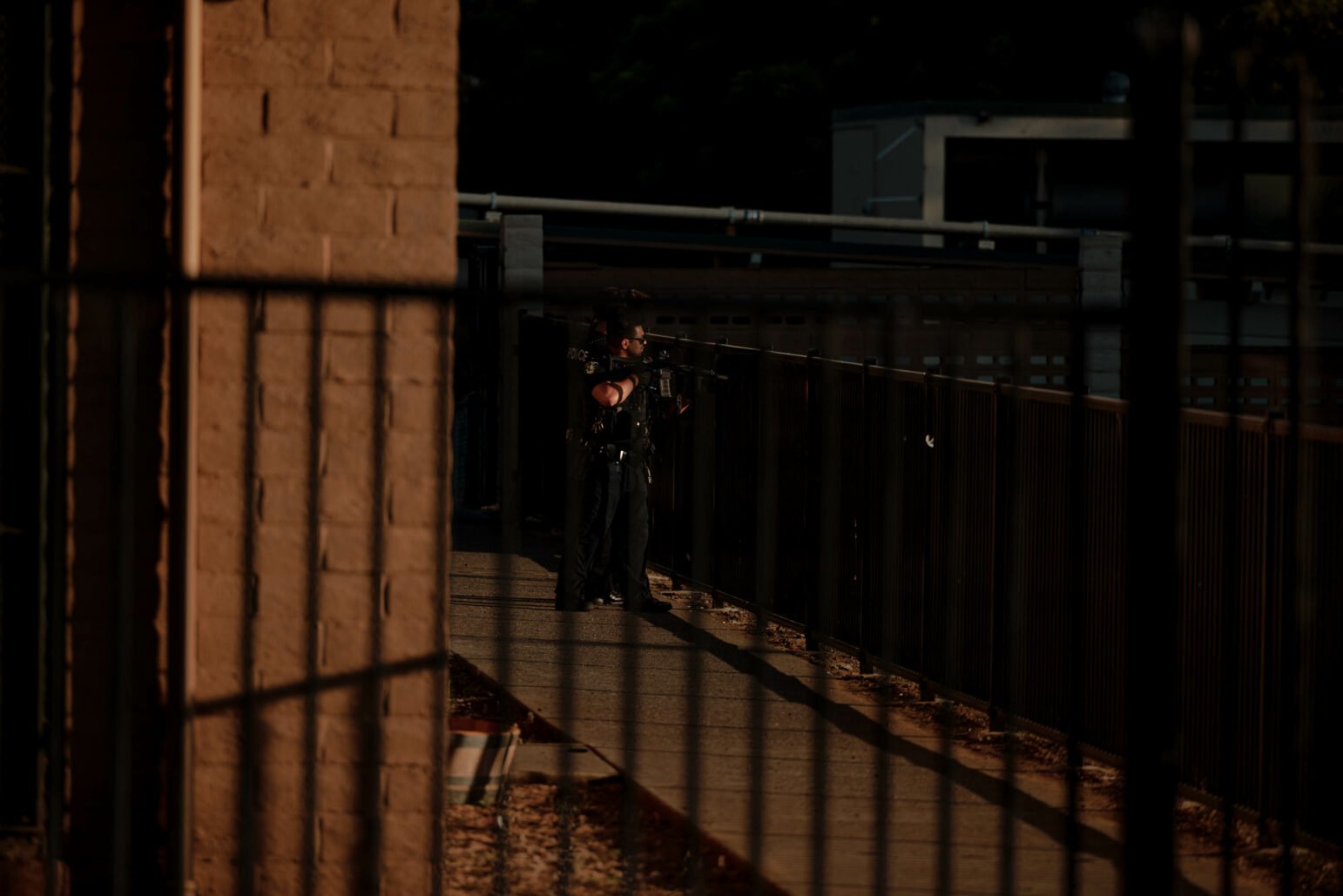 Two police officers with rifles stand near a fence outside a building, surveying the area.