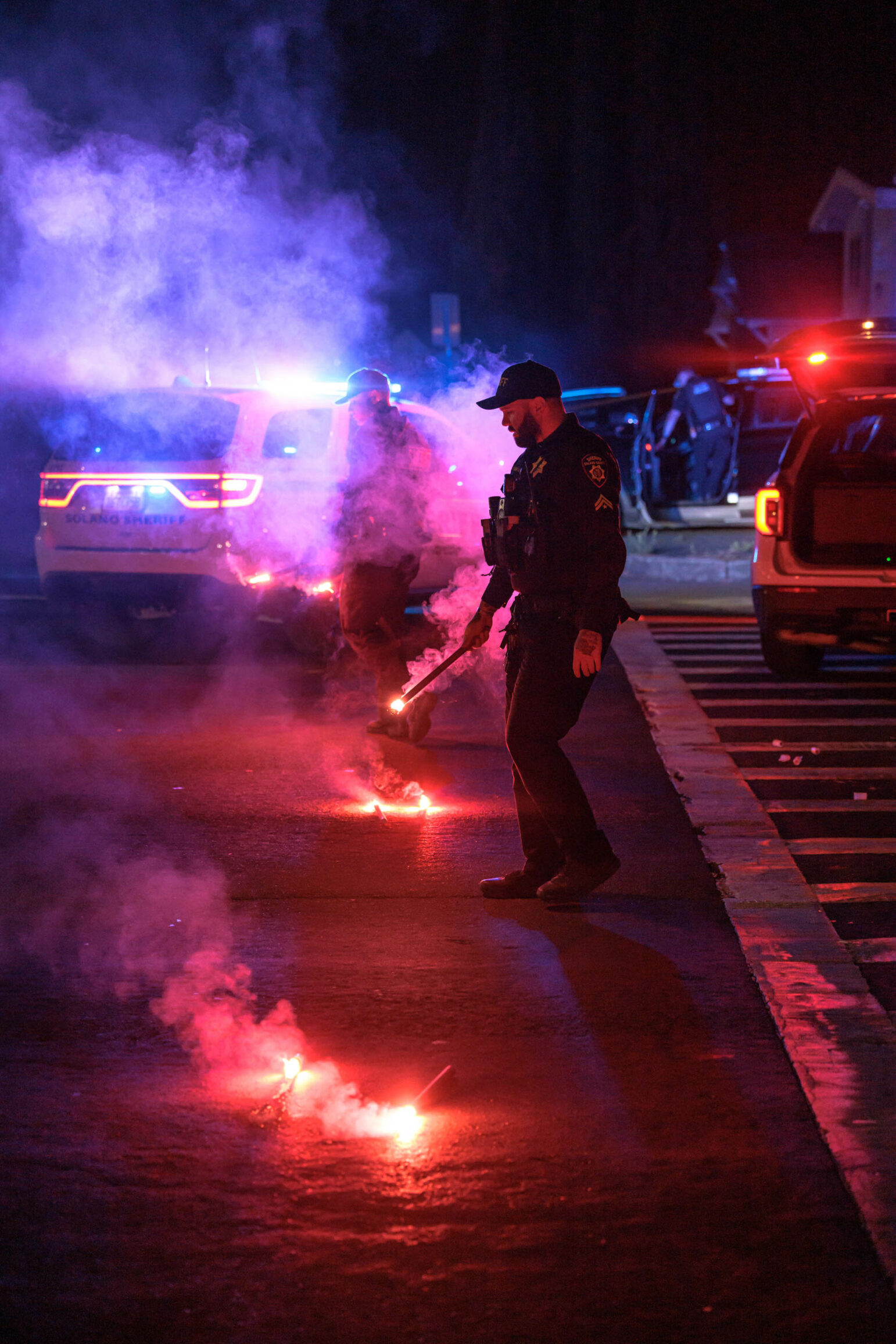 Sheriff's deputies handling flares at the scene of an incident at night, with bright red and blue lights illuminating the area.