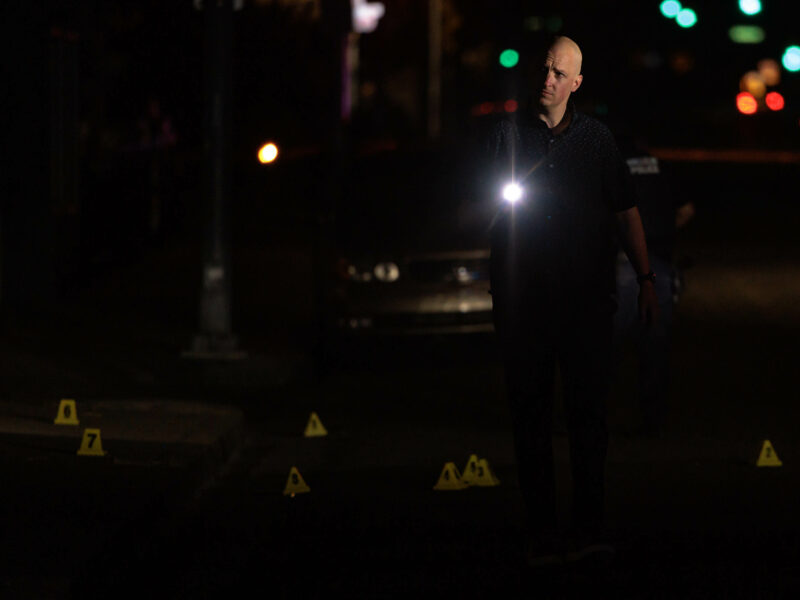 A police investigator walking in the dark, holding a flashlight that illuminates evidence markers on the ground.