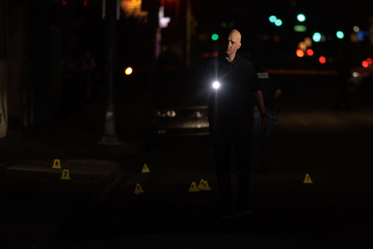 A police investigator walking in the dark, holding a flashlight that illuminates evidence markers on the ground.