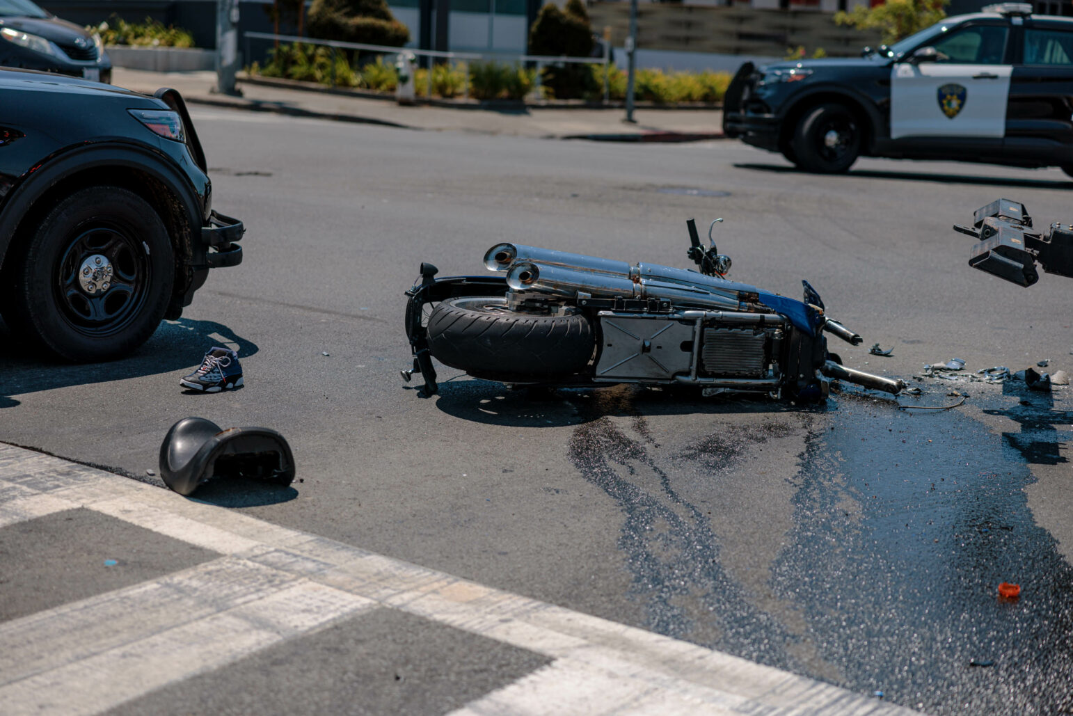 A destroyed motorcycle lays in the road with fluid leaking from it with Vallejo police SUVs in the background during daytime.