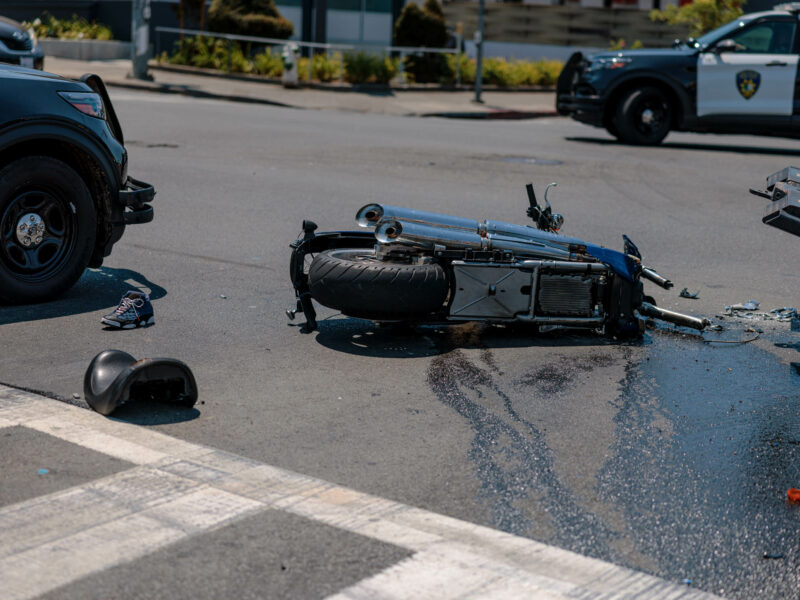 A destroyed motorcycle lays in the road with fluid leaking from it with Vallejo police SUVs in the background during daytime.