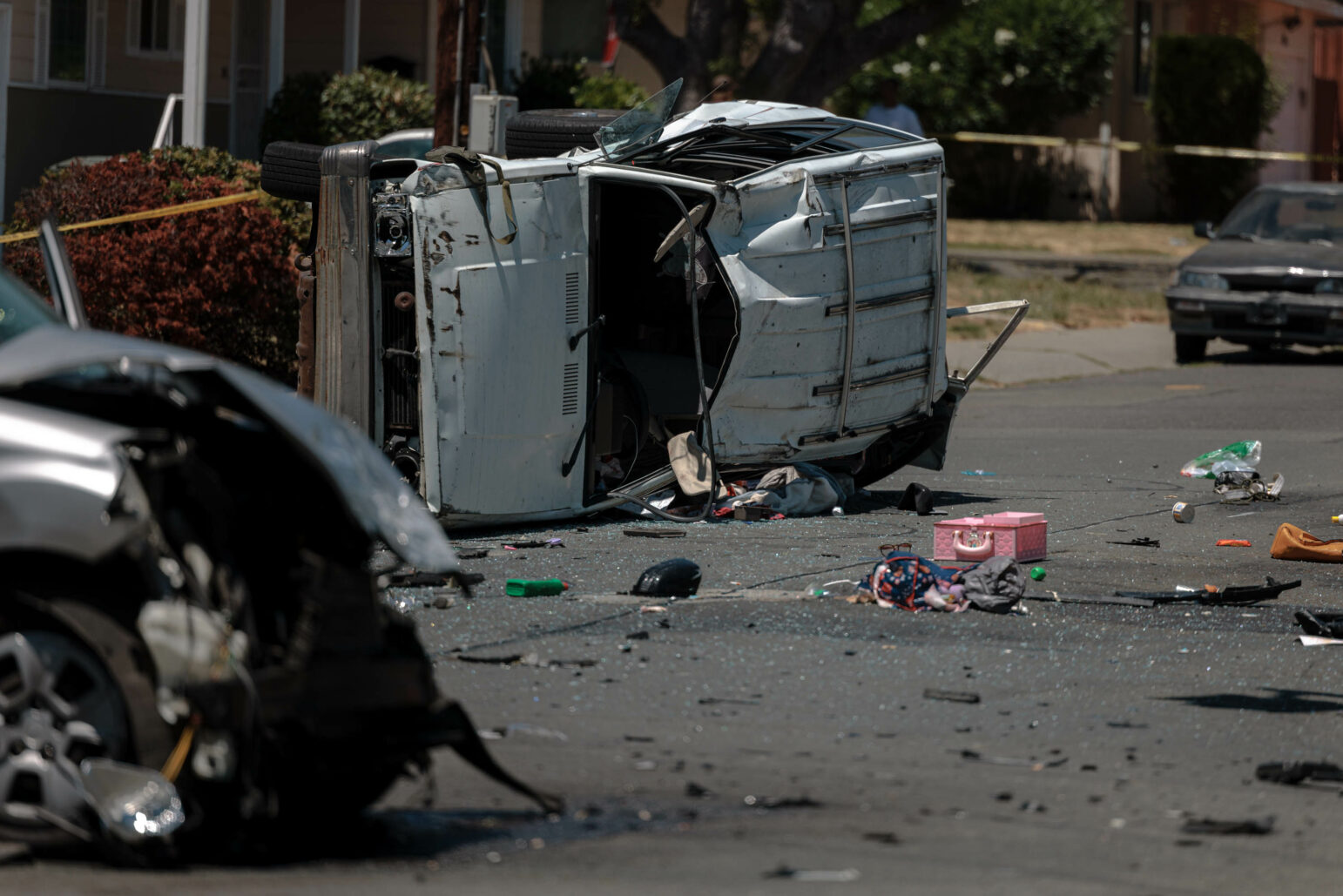 A Isuzu Trooper lies on its side in the middle of a street after a collision, surrounded by debris including car parts and personal items like a pink box and clothing.