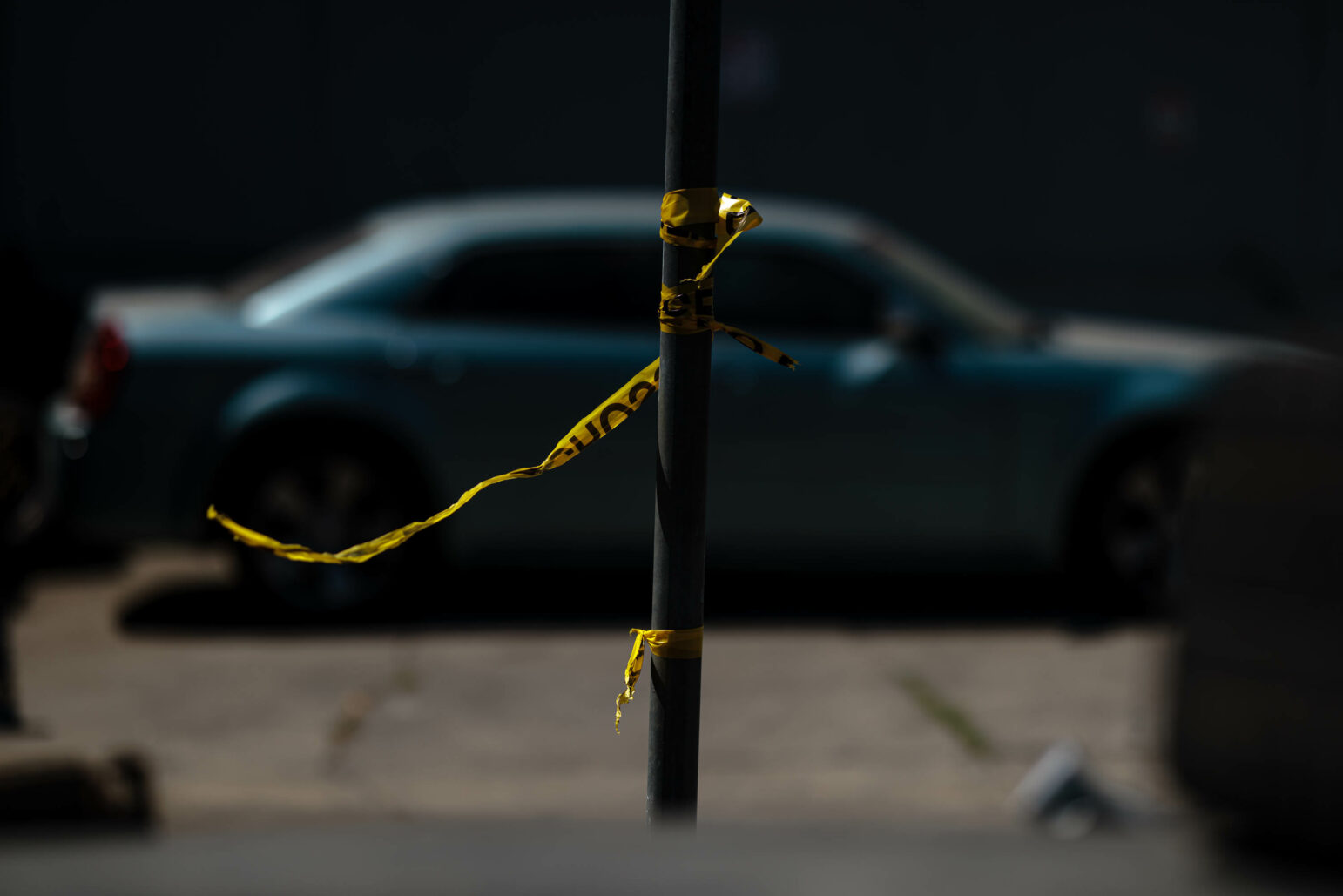 Police tape tied to a pole blows in the wind during daytime. A blue car is visible, out of focus, in the background.