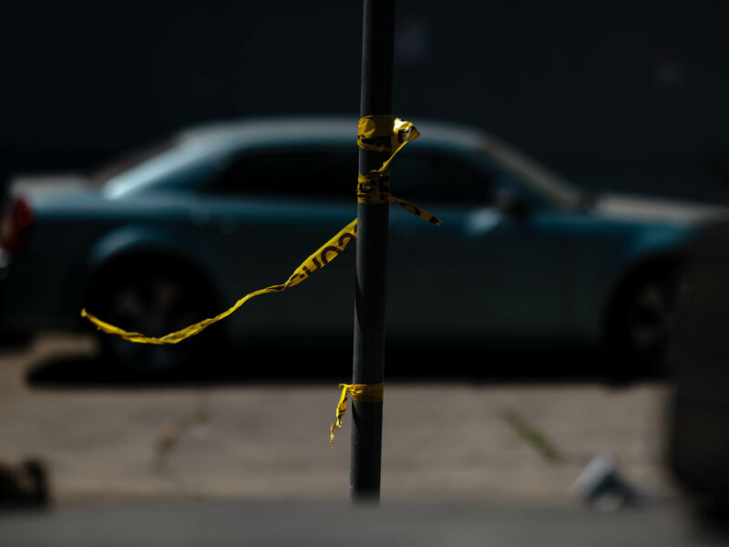 Police tape tied to a pole blows in the wind during daytime. A blue car is visible, out of focus, in the background.