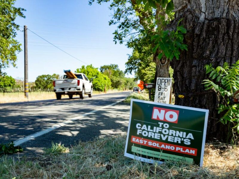 A protest sign against the "California Forever" project is seen in the foreground, reading "NO to California Forever's East Solano Plan." The sign is placed on the grass beside a road, near a large tree. In the background, a white pickup truck is driving away, and there is greenery and trees lining the roadside.