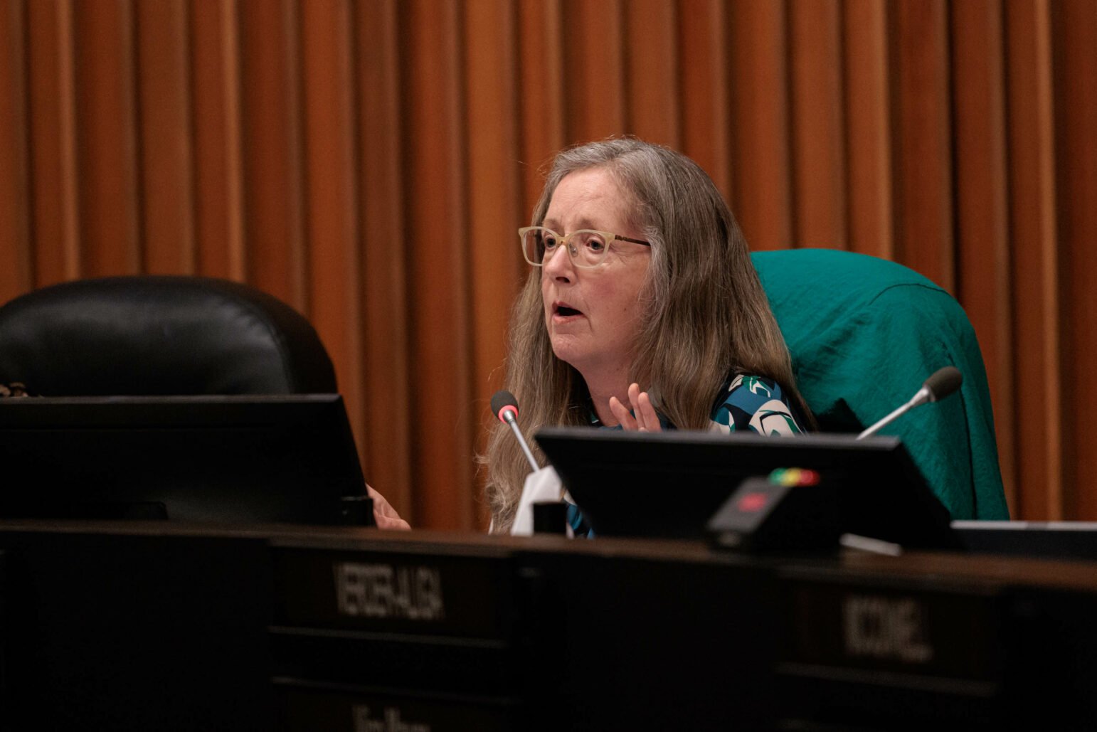 A councilwoman speaks at a podium during a council meeting. She is middle-aged with long grey hair and glasses, gesturing with her hands as she addresses the chamber, which is characterized by wooden interiors and formal seating.