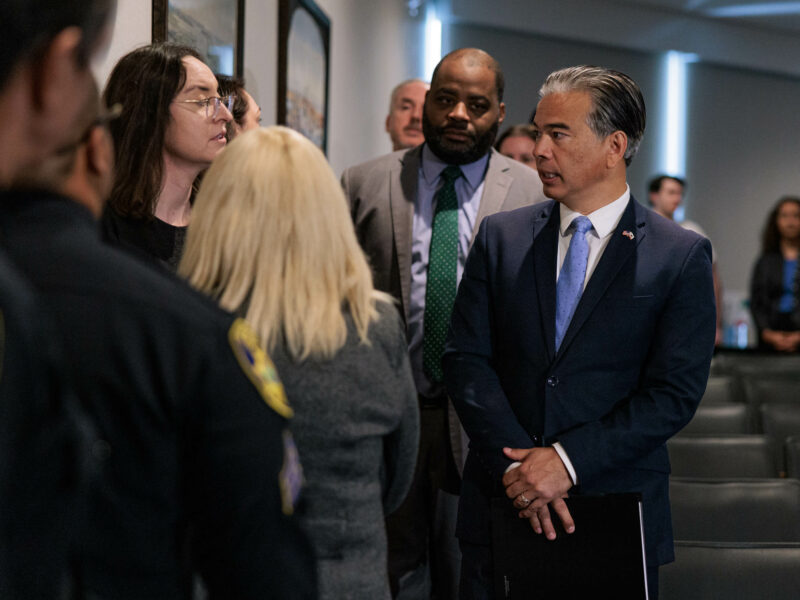 California Attorney General Rob Bonta prepares to speak at an indoor event. He is surrounded by other attendees and attorneys, some in conversation, within a city council chambers adorned with framed pictures on the walls.