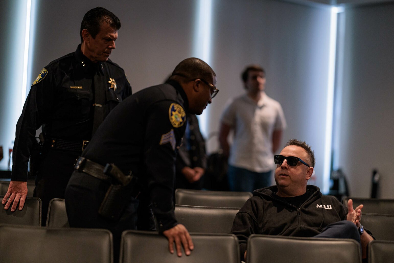 Two police officers converse with a seated man wearing sunglasses and a black hoodie inside a city council chambers. The focus is on their interaction with other individuals blurred in the background.