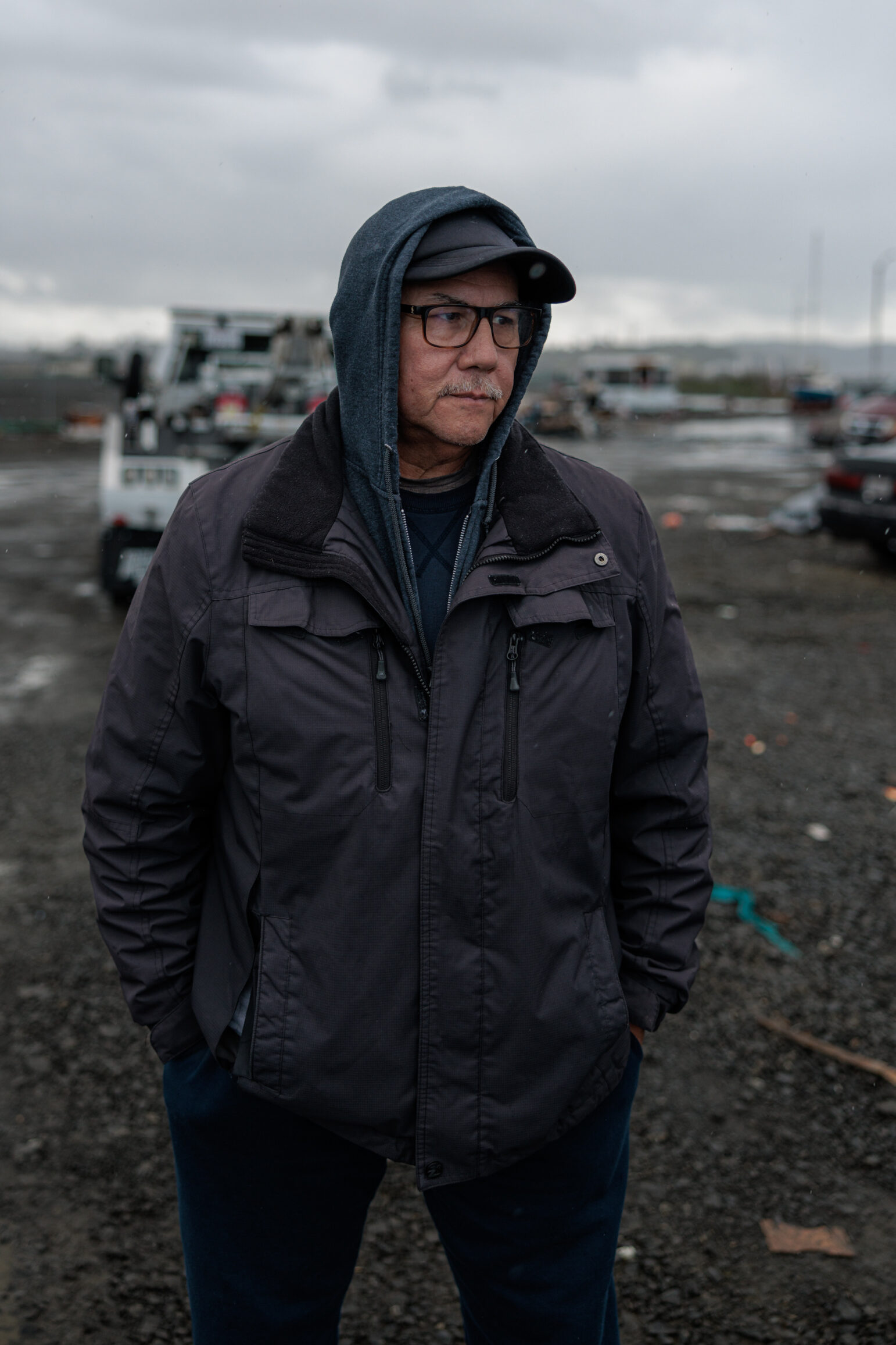 A person in a hooded jacket and glasses stands in a homeless encampment, with debris and vehicles in the background. The sky is overcast, and the ground is muddy.