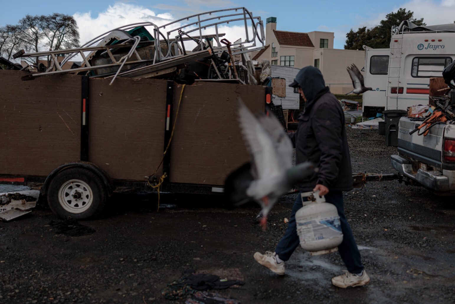 A person dressed in a hooded jacket and cap is walking with a propane tank in hand. Pigeons are flying around, and in the background, there is a trailer filled with various metal objects.