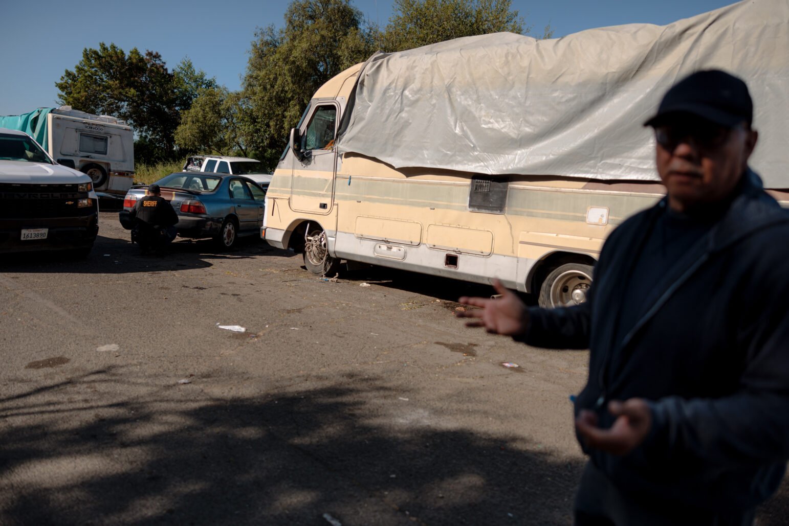 A sheriff's deputy checks an apparently abandoned car near a homeless encampment. A person in the foreground is gesturing toward the scene.