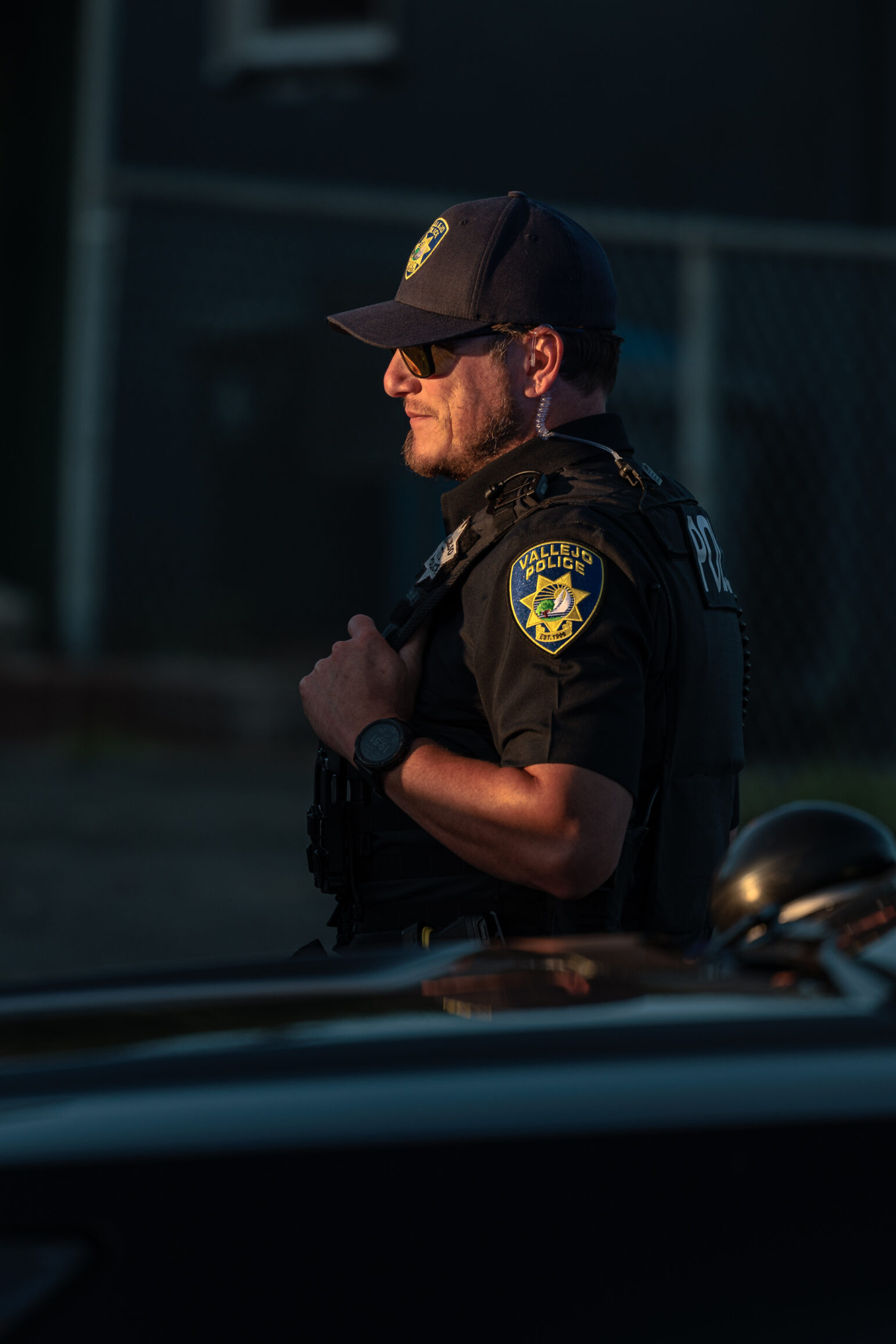 A close-up of a Vallejo police officer wearing sunglasses, a dark uniform, and a cap. The officer is standing outdoors with sunlight illuminating part of the scene.