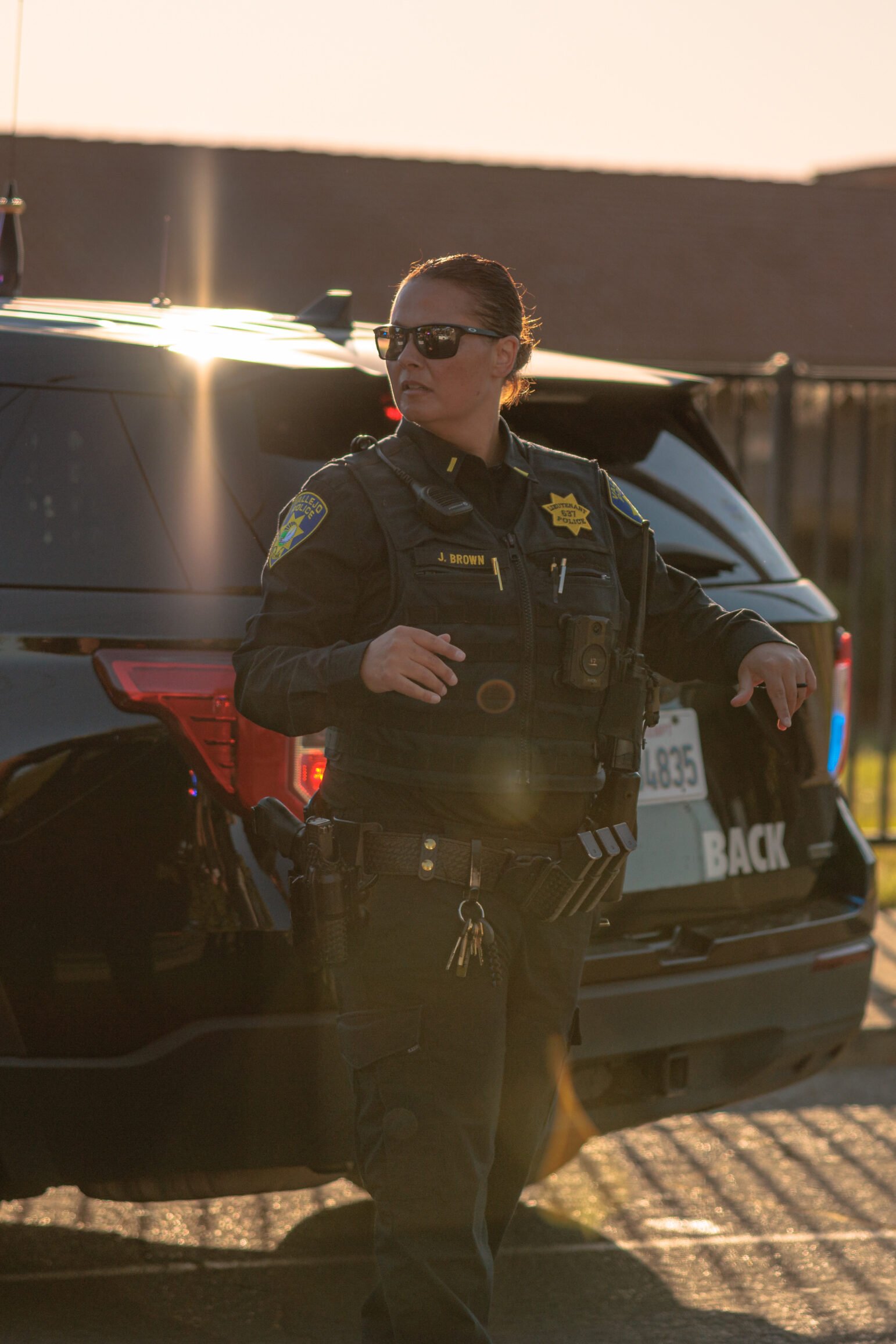 A Vallejo police officer, wearing a vest and sunglasses, stands next to a police vehicle with the setting sun casting a warm glow. The officer appears to be observing something off-camera.