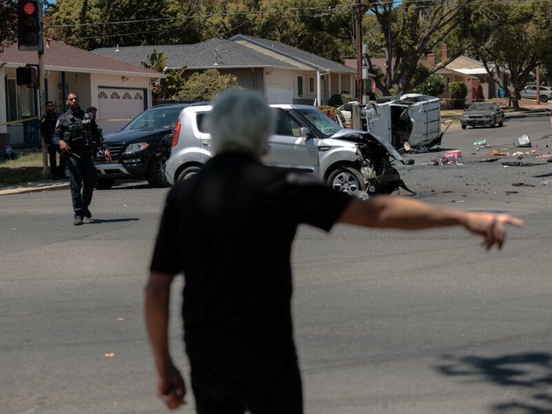 A man points near a street scene of a severe vehicle collision, where cars are visibly damaged and debris is scattered across the road. The focus includes officers surveying the scene and securing the area in a suburban neighborhood under clear skies.