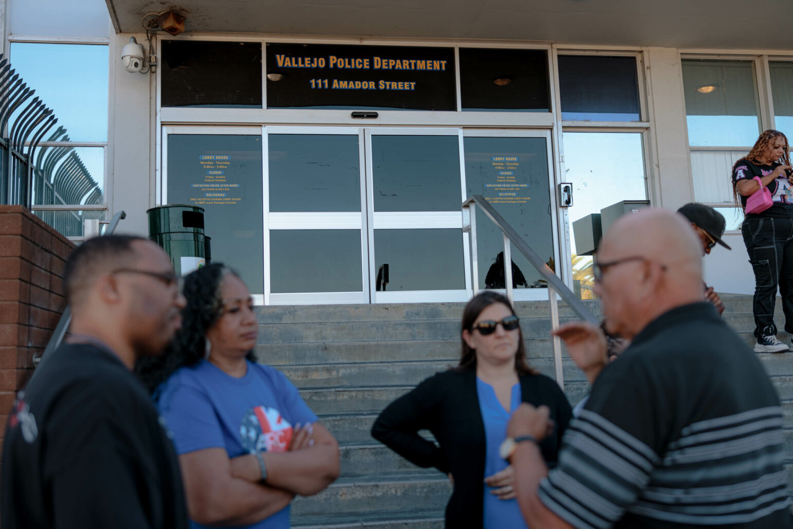 A group of people gather outside the Vallejo Police Department at 111 Amador Street. Several individuals are shown in the foreground, including a woman in a blue shirt and a man in black, both with contemplative expressions. The building's entrance features signage displaying the address and office hours, with more people visible on the steps.