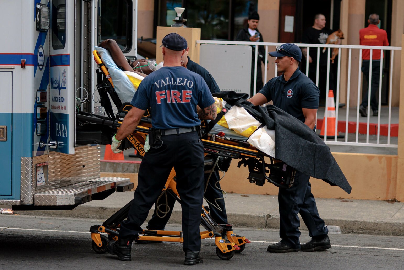 Emergency responders from the Vallejo Fire Department and an ambulance company are pictured loading a patient on a stretcher into an ambulance. The patient is covered with a yellow blanket and surrounded by medical equipment, highlighting the urgency of the situation. The scene is set in front of a building, with bystanders in the background, underscoring the public nature of the emergency.