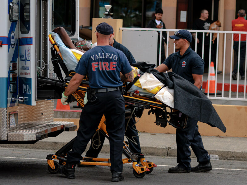 Emergency responders from the Vallejo Fire Department and an ambulance company are pictured loading a patient on a stretcher into an ambulance. The patient is covered with a yellow blanket and surrounded by medical equipment, highlighting the urgency of the situation. The scene is set in front of a building, with bystanders in the background, underscoring the public nature of the emergency.