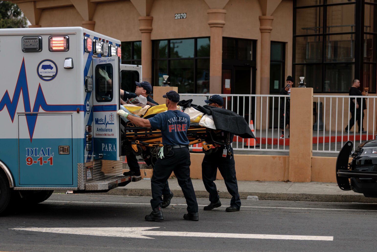 Emergency medical responders and firefighters load a patient on a stretcher into an ambulance outside a terracotta-colored building during daytime.