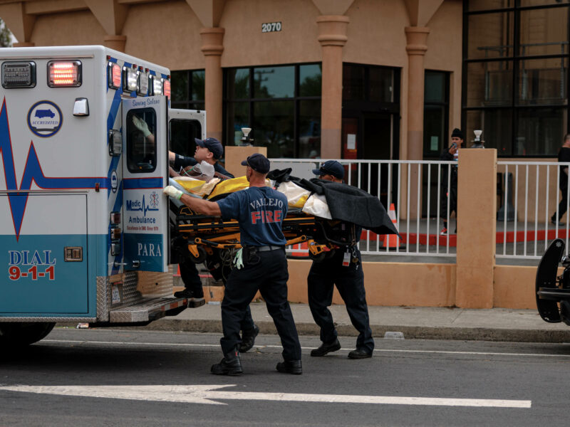 Emergency medical responders and firefighters load a patient on a stretcher into an ambulance outside a terracotta-colored building during daytime.