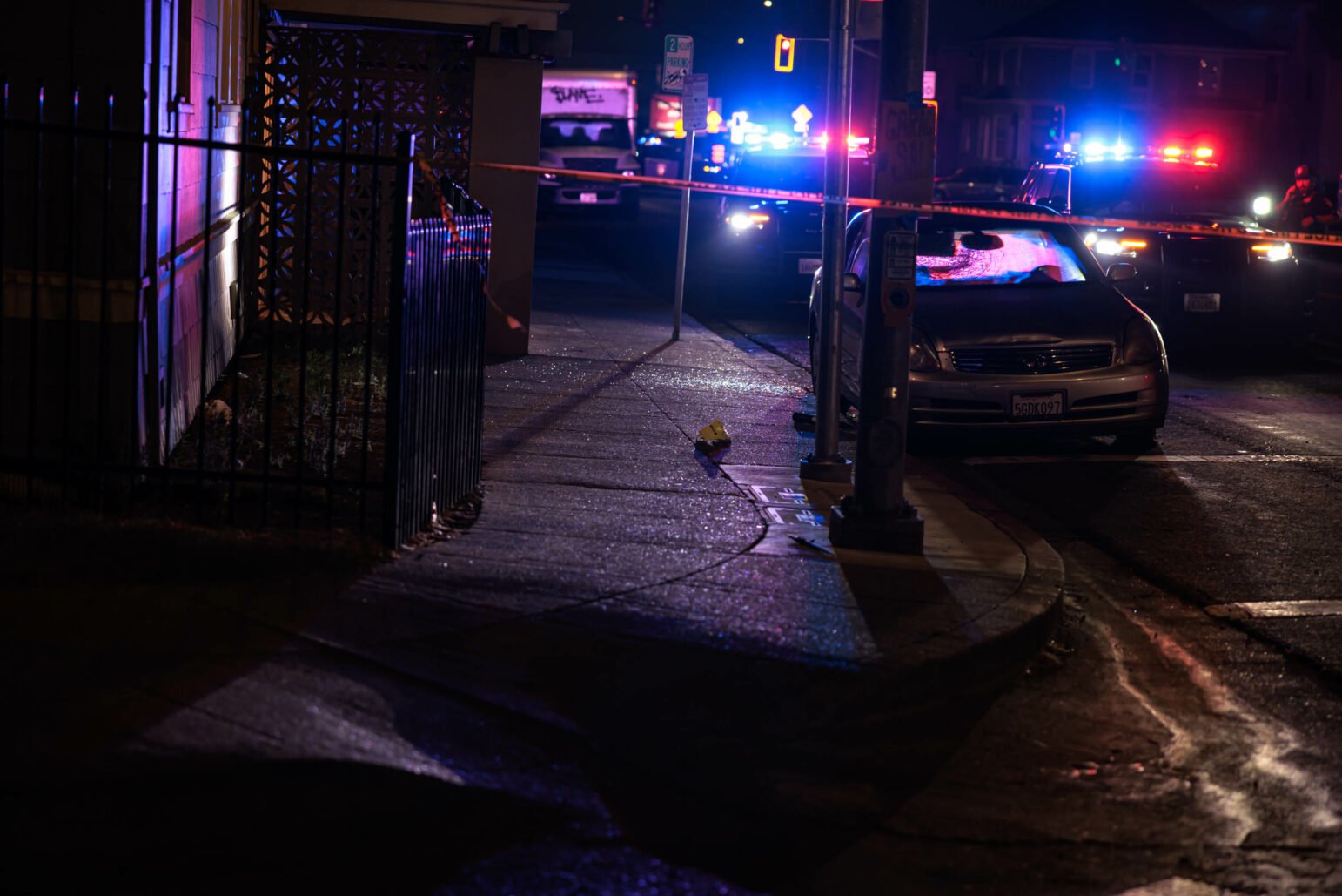 A nighttime scene on a suburban street. Police vehicles display red and blue flashinging lights in the background. The foreground shows crime scene tape and, behind that, a car with a shattered window.